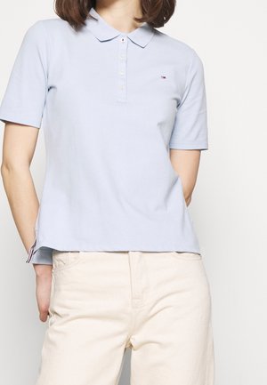 Femme portant un polo bleu clair à manches courtes avec un petit logo et un pantalon couleur crème sur fond uni.