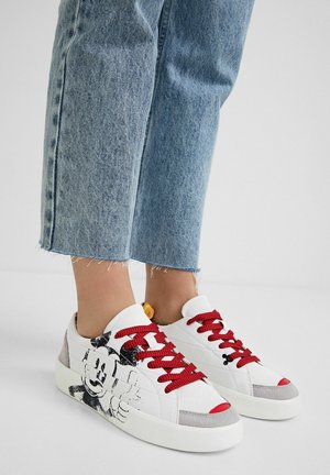 Personne portant un jean court bleu clair et des baskets blanches avec des lacets rouges et un dessin de personnage de dessin animé noir et blanc sur le côté.