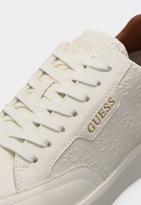 Gros plan sur une basket blanche Guess avec un motif embossé et le logo Guess doré sur le côté, avec des lacets blancs et des détails de couture.