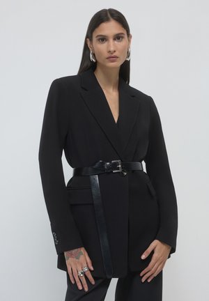 Black tailored blazer with a deep V-neck, structured shoulders, and a glossy leather belt. Features side pockets and sleek buttons.
