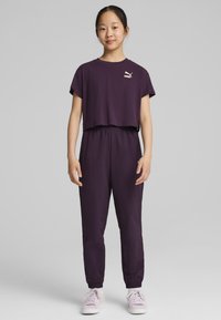 Violet trumpas marškinėliai su mažu baltu logotipu; atitinkamos violetinės joggingo kelnės su elastinga juosmena; šviesiai violetiniai sportbačiai.