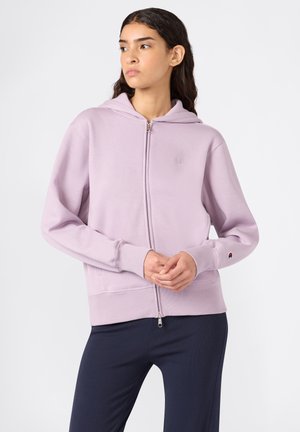 Femme portant un sweat à capuche zippé de couleur violet clair, se tenant avec les mains jointes, regardant sur le côté, associée à un pantalon sombre.