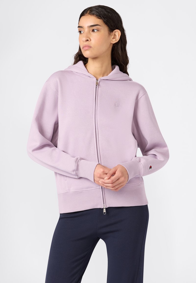 Donna che indossa una felpa leggera viola con zip e cappuccio, in piedi con le mani unite, che guarda di lato, abbinata a pantaloni scuri.