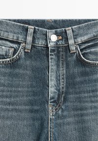 Primo piano della vita di jeans di denim blu, con bottone, passanti per cintura e tasche frontali con dettagli di cuciture visibili.