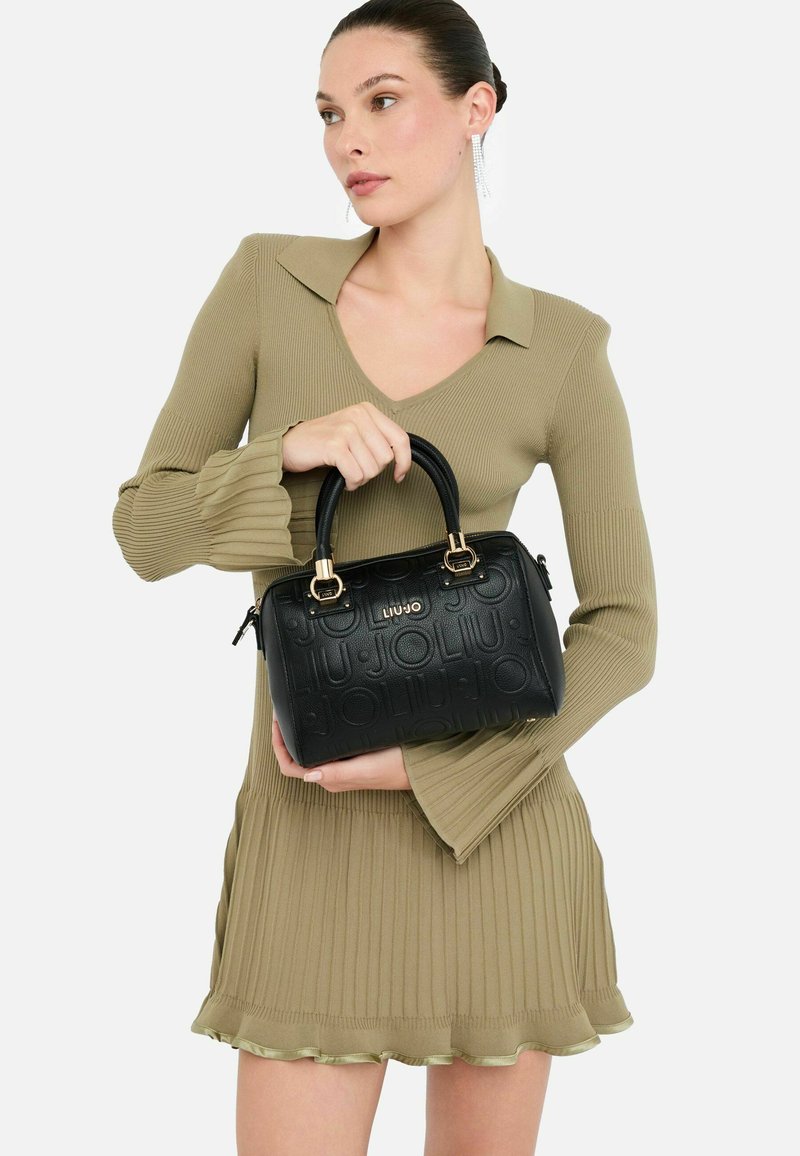 Sac à main en cuir noir avec motif embossé, poignées supérieures doubles, quincaillerie dorée et fermeture éclair. Le mannequin porte une robe moulante à côtes olive.