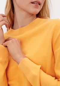Gul strikket sweater med ribbet rund halsudskæring og lange ærmer. Stoffet ser blødt ud med en glat tekstur. Close-up af hænder, der justerer halsudskæringen.