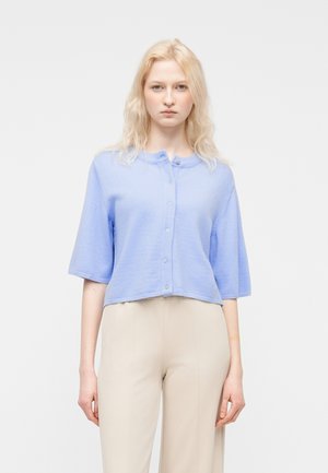 Mujer joven con un cárdigan azul claro de manga corta abotonado y pantalones beige de talle alto, de pie frente a un fondo blanco liso.