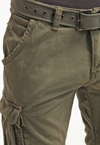 Pantalones cargo en tela verde oliva, con múltiples bolsillos, un cinturón texturizado con hebilla de metal y un diseño de ajuste relajado.
