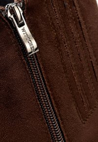 Botín de ante marrón con una cremallera metálica que lleva grabado el nombre de la marca, costuras de acento y una superficie texturizada.