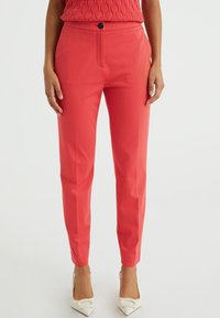 Woman wearing salmon pink fitted trousers with a black button and white pointed heels, standing against a white background.