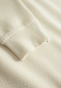 Fehér, textúrált szövet, amely vaffel-kötött mintával rendelkezik, és azonos színű bordázott mandzsetta díszíti. A hangsúly a szövet textúráján és a tervezési részleteken van.