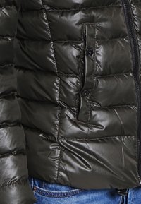 Doudoune verte foncée, avec un extérieur en nylon brillant, un motif matelassé, une poche avant à bouton et une fermeture éclair.