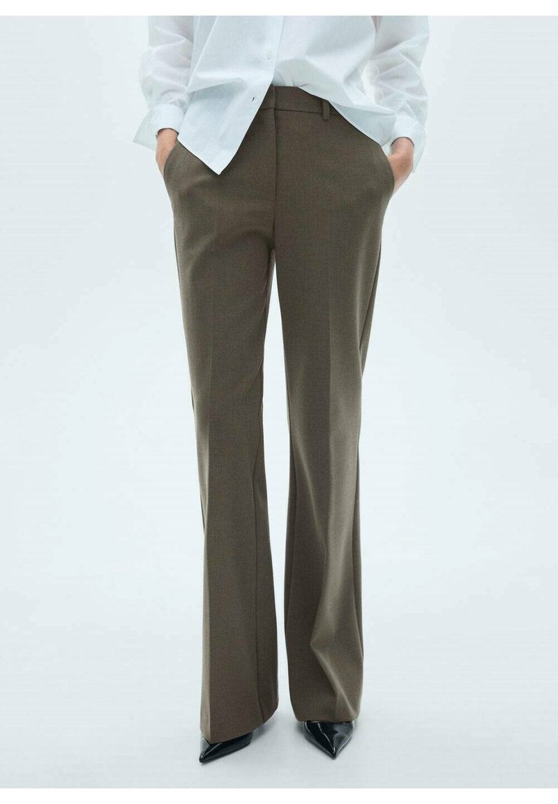 Personne portant un pantalon marron à jambes larges, une chemise blanche boutonnée, les mains dans les poches, et des chaussures noires à bout pointu, debout sur un fond blanc.