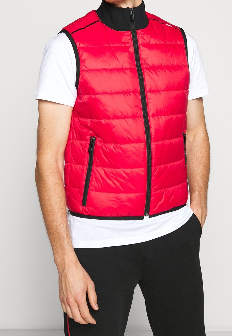 Homme portant un gilet matelassé rouge sans manches avec des fermetures éclair noires par-dessus un t-shirt blanc à manches courtes et un pantalon noir, debout avec une main dans la poche.