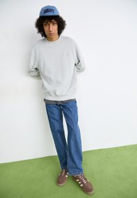Grå sweatshirt, blå kasket, løse denim jeans i en lys vask, og brune sneakers med hvide striber, præsenteret mod en ensfarvet baggrund.