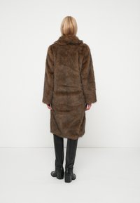 Brūns mākslīgā pūka mētelis ar plašu apkakli, garām piedurknēm un brīvu piegriezumu, kas sasniedz vidus teļu. Valkāts virs melnajiem ādas ceļgala zābakiem.