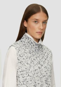 Pleteni bezrukavni turtleneck u bijeloj boji s crnim točkicama. Ima visoki ovratnik i teksturiranu tkaninu, složen preko bijele košulje.