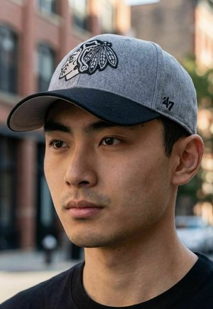 Young man wearing gray and black cap with a Native American chief logo, standing outdoors in an urban setting.