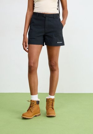 Persoon draagt zwarte Columbia-shorts, witte sokken, bruine veterlaarzen en een wit top, staand op een groene vloer tegen een witte achtergrond.