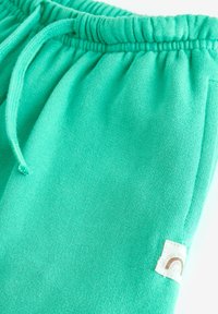 Turquoise joggingbroek met een elastische tailleband, trekkoord en een klein stoffen label met een bruin boogontwerp aan de zijkant.