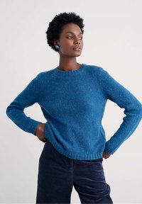 Rövid göndör hajú nő, aki kék pulóvert és sötét kordbársony nadrágot visel, keze csípőn állva, az oldalra néz.