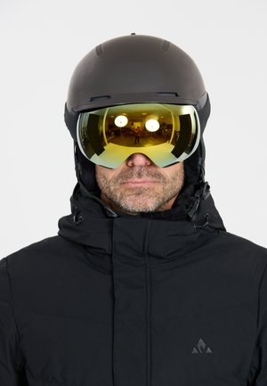 Man wearing black ski helmet, reflective yellow ski goggles, and black winter jacket with logo on chest, facing forward against white background.