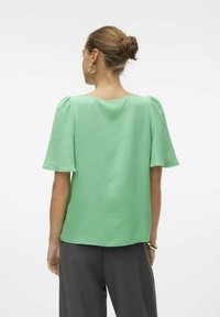 Top a maniche corte in tessuto verde chiaro texturizzato, con scollo rotondo e vestibilità rilassata, abbinato a pantaloni grigi larghi.