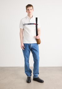 Hvit polo med horisontale striper i marineblått, rødt og svart, med "TOMMY HILFIGER" logo. Sammen med blå denim jeans og svarte sko.