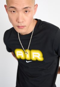 Svart t-skjorte med korte ermer som har en fremtredende gul logo som staver "AIR" og en liten hvit Nike swoosh. Sølvkjede tilbehør.