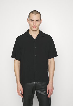 Chemise noire à manches courtes et à col camp, fabriquée en tissu léger. Dispose de boutons discrets pour la fermeture.