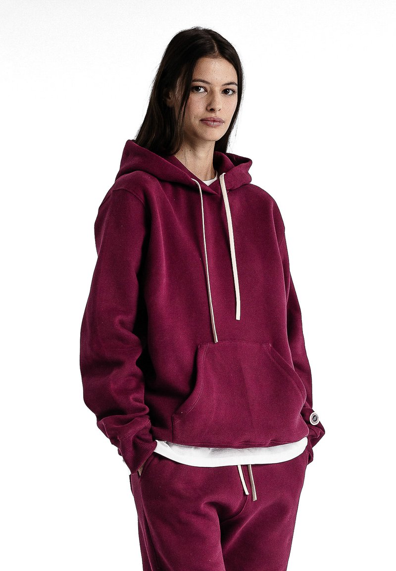 Un sweat à capuche bordeaux avec cordons de serrage, poche avant et poignets côtelés. Stylisé avec une chemise blanche en superposition en dessous et un pantalon bordeaux assorti.