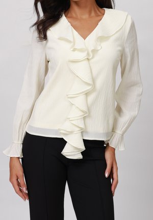 Frau trägt eine weiße Bluse mit langen Ärmeln, gerafftem Vorderteil und Manschetten, kombiniert mit schwarzen, eng anliegenden Hosen vor einem schlichten Hintergrund.