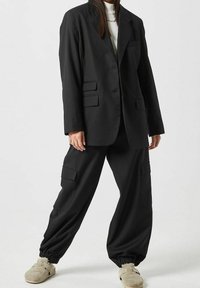 Zwarte oversized blazer met twee voorzakken en bijpassende zwarte cargobroek met elastische boorden, gecombineerd met beige instapschoenen.