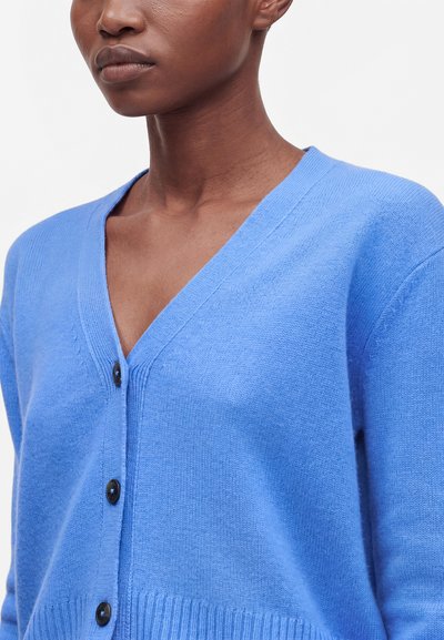 Femme portant un cardigan bleu clair à col en V avec des boutons noirs, montrée des épaules à la poitrine sur un fond uni.