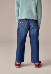 Vaqueros de mezclilla con un corte relajado, color azul, con bordados florales en los bolsillos traseros. Combinados con zapatos rojos y rosas.