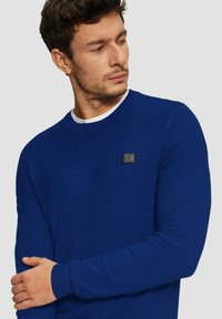 Blauer strukturierter Pullover mit Rundhalsausschnitt, gerippten Bündchen und einem schwarzen Logo-Patch auf der Brust. Glatte Strickqualität.