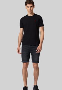 Czarny t-shirt z okrągłym dekoltem i czerwonym logo na piersi, w zestawieniu z ciemnymi dżinsowymi szortami podwiniętymi na końcu. Model w czarnych trampkach.