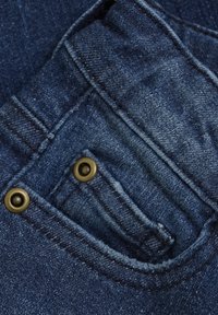 Denimtyg i mörkblått med en liten framficka som har två gyllene nitar. Synlig sömnad och texturdetaljer förstärker den övergripande designen.