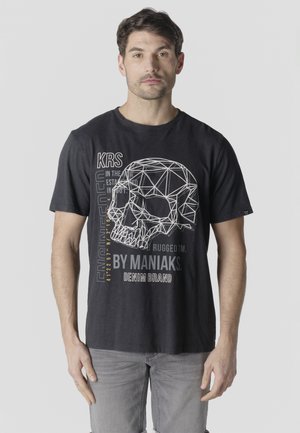 Man draagt een zwart T-shirt met een witte geometrische schedelafbeelding en tekst, gecombineerd met lichtgrijze jeans, staand tegen een eenvoudige achtergrond.