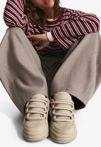 Bézs színű tornacipők lekerekített orral, texturált anyagból és feltűnő fűzőkkel. Lazán illeszkedő, világosszürke, bőszárú nadrággal és bordó csíkos felsővel párosítva.