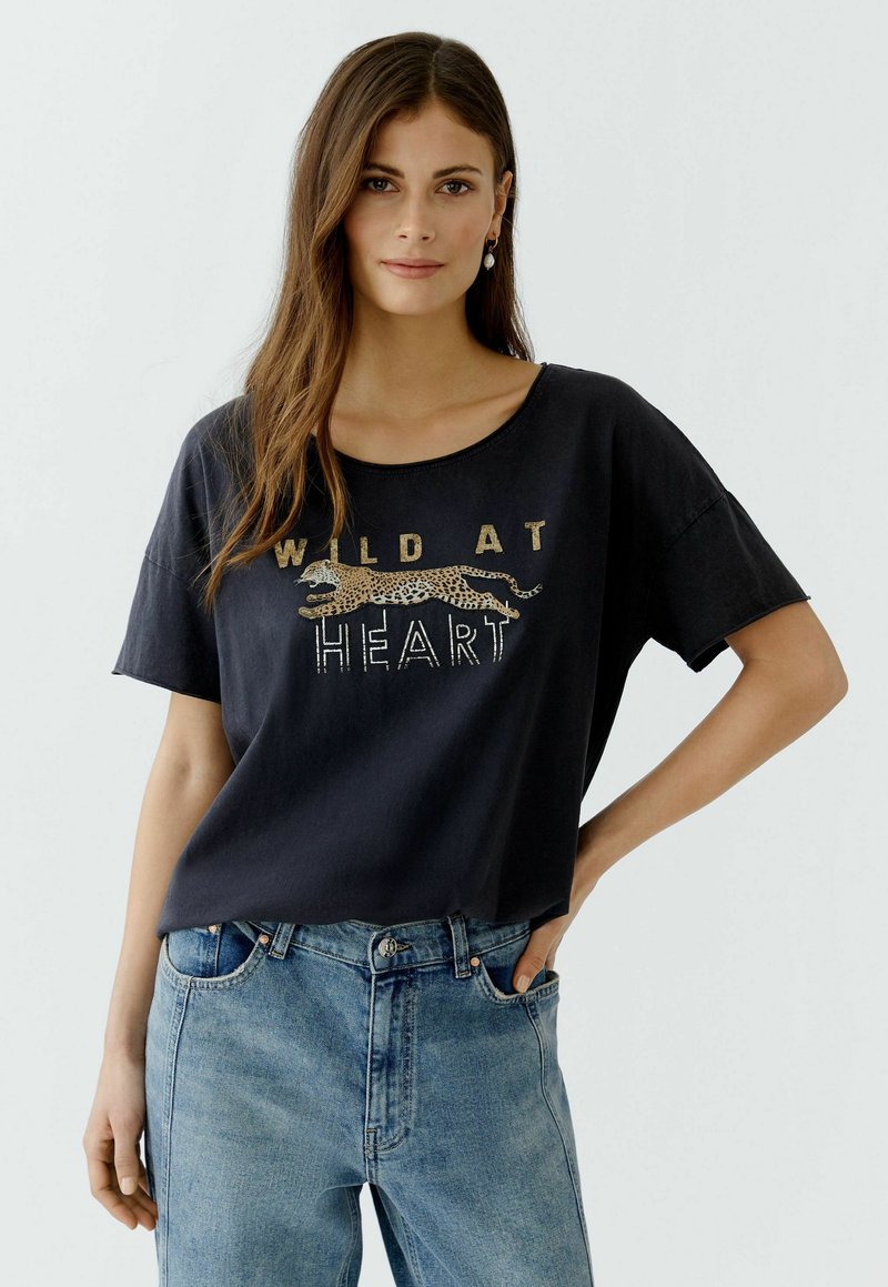 Black cotton T-shirt with a relaxed fit, featuring gold text "WILD AT HEART" and a leopard illustration. Paired with light blue high-waisted jeans.