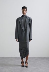 Pelēks pielāgots žakets virs atbilstoša turtleneck un pieguļošām svārkiem. Komplektā ir drapēts dizains un norādītas papēžu kurpes.