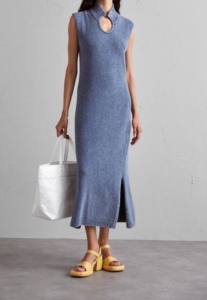 Kobieta w niebieskiej sukience dzianinowej bez rękawów z rozcięciem z przodu, trzymająca białą torbę typu tote i żółte sandały na platformie na tle szarej ściany.