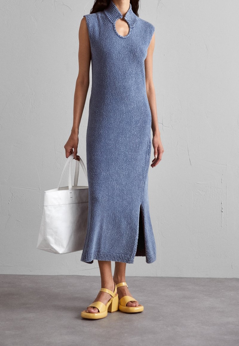 Femme portant une robe en maille bleue sans manches avec une fente à l'avant, tenant un sac fourre-tout blanc et portant des sandales compensées jaunes, devant un mur gris.