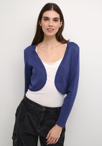 Blue cropped cardigan made of soft material with long sleeves, rounded edges, and a smooth texture, worn over a white top.