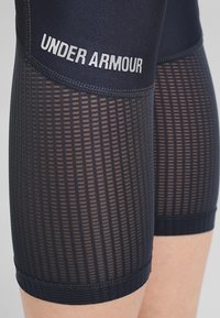 Laadun verkkosivut kengät, joissa on hengittävä verkkotekstuuri, sileä paneelointi ja "UNDER ARMOUR" painettuna vyötärölle.