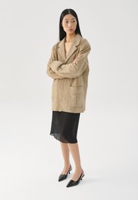 Beige semišová oversized bunda se dvěma předními kapsami, nošená přes černou plisovanou sukni, doplněná černými lodičkami na podpatku s špičatou špičkou.