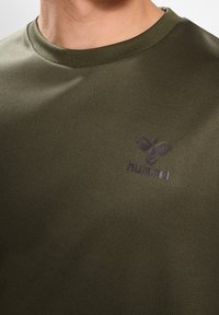 Närbild av en olivgrön t-shirt med rund hals och svart hummel-logotyp på bröstet, bärs av en person som visas från halsen och neråt.