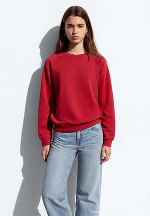 Junge Frau steht an einer weißen Wand, trägt einen roten Sweatshirt mit langen Ärmeln und hellblaue Jeans, Ohrringe und einen neutralen Gesichtsausdruck.
