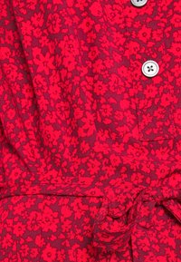 Tissu de robe fleurie rouge avec de petites fleurs rouges vives sur un fond rouge profond, avec une taille nouée et des détails de bouton.